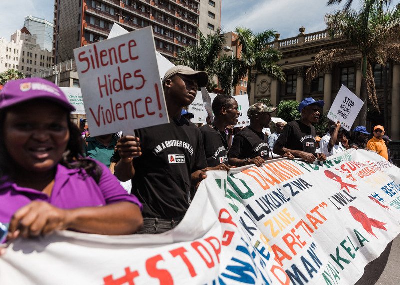 People marching holding a large protest banner and a placard that says: 'silence hides violence!'.