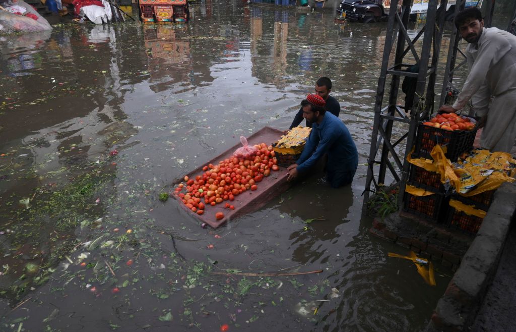 Men stand in high flood waters, directing a wooden cart loaded with tomatoes. 