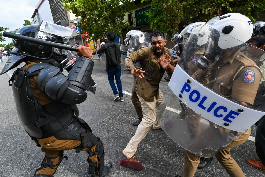 Heavily armed police raise baton towards cowering protester.