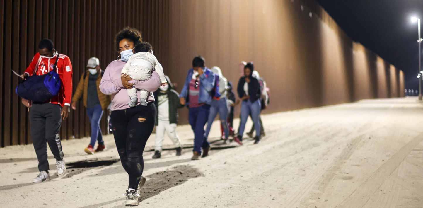 Immigrants walk along the U.S.-Mexico border barrier in the early morning hours under the light of street lamps