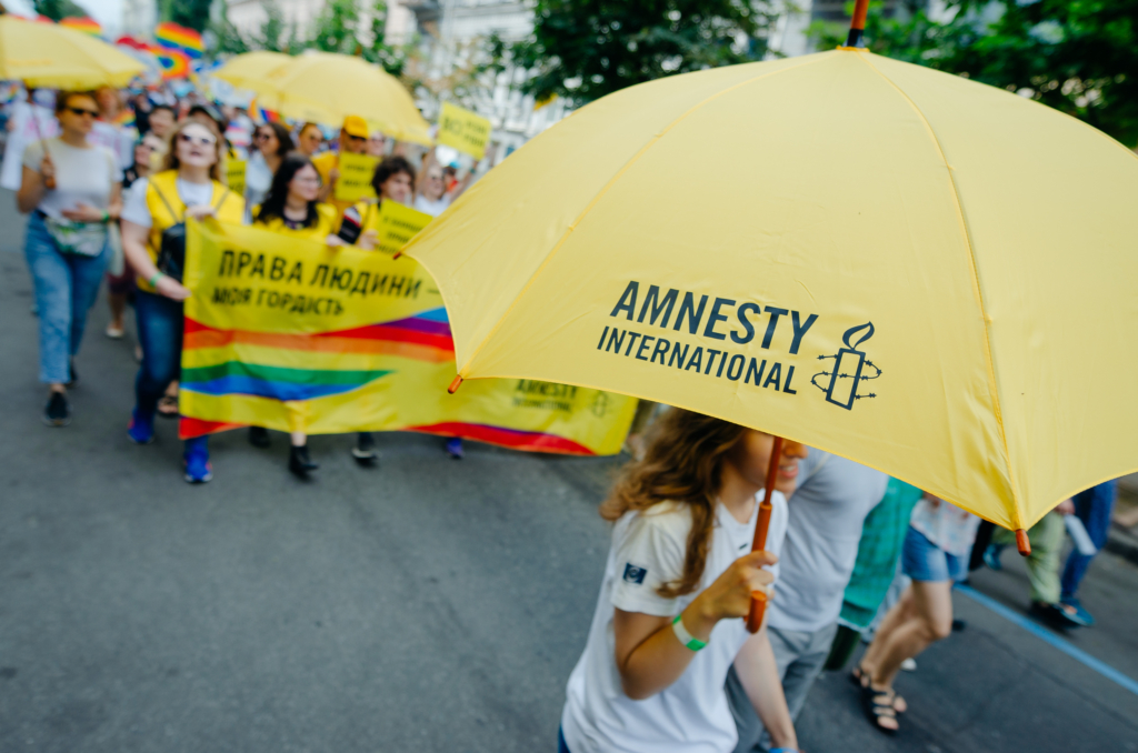 someone is holding a yellow umbrella with the Amnesty International logo on it. They are walking in front of a Pride March in Kyiv. 