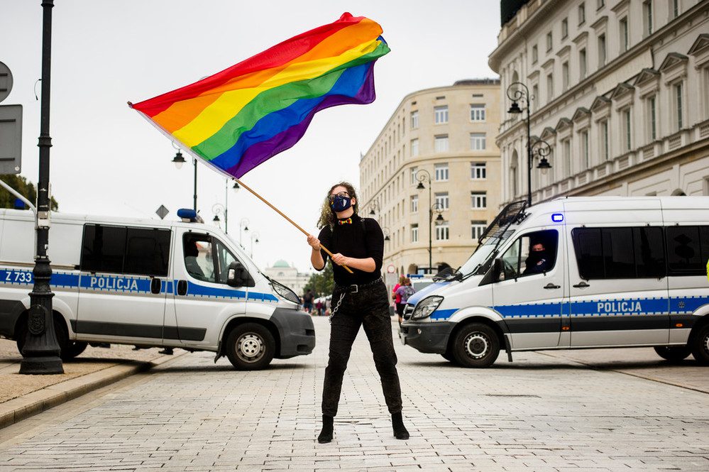 someone is waving a rainbow flag in the air. Behind them sit two large police vans.