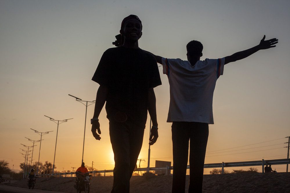 Two people are backlit by a sunset. One of them is holding their arms out wide. 