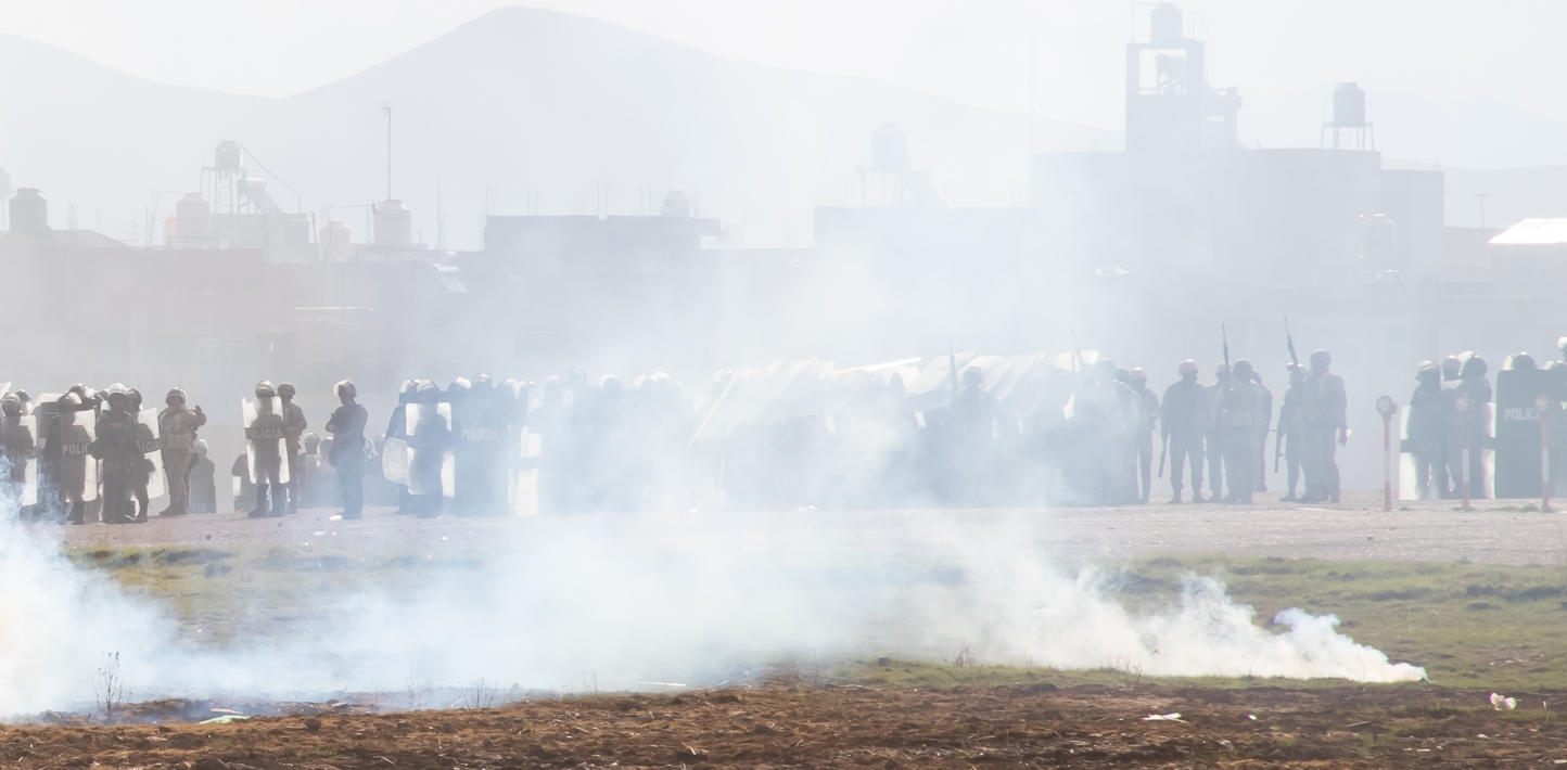 Heavily armed security personnel stand in a line, partially obscured by the smoke from a tear gas cannister.