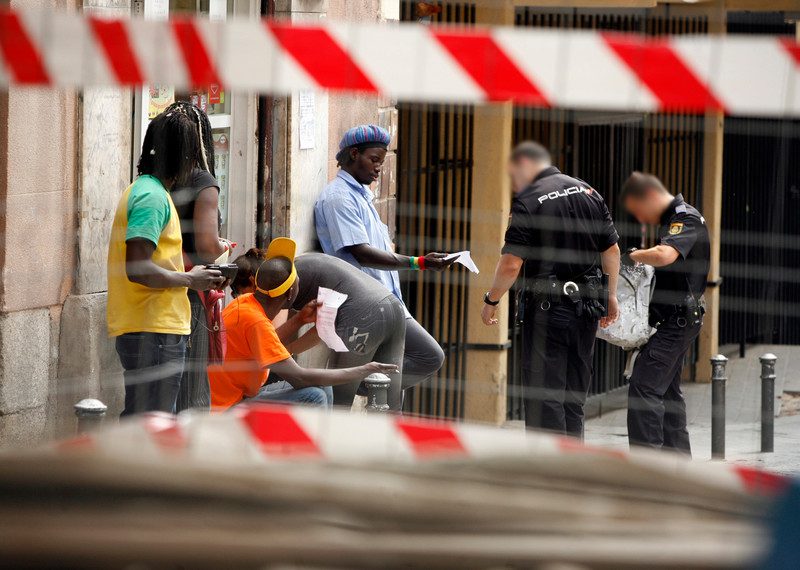 Through a fence, two police officers are seen searching a group of five black people, some of whom are holding out papers.