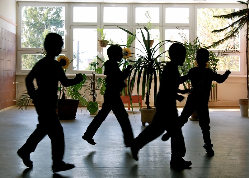 Siluetas de niños jugando en una gran sala de escuela con plantas en el fondo.
