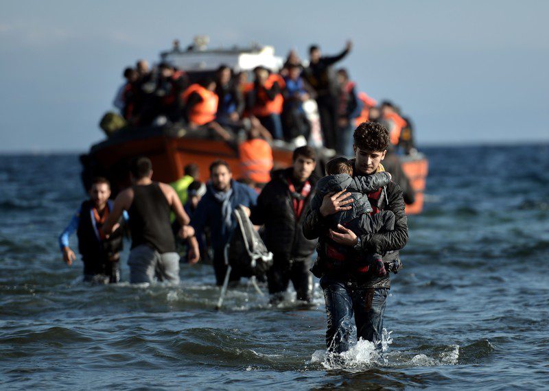 A crowded boat in the sea, with people climbing off and wading through the water. A small child is carried in the foreground.