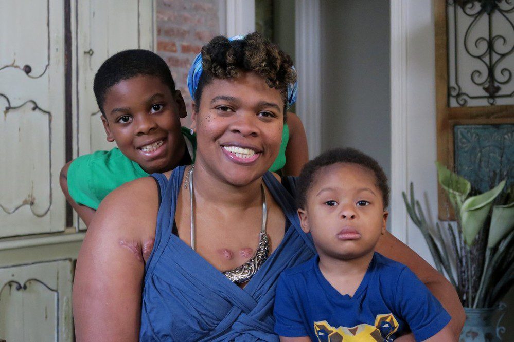 Angela, a survivor of gun and domestic violence sits with her two young sons. The trio are smiling, but you can see visible scars on Angela’s chest that were the result of domestic violence.