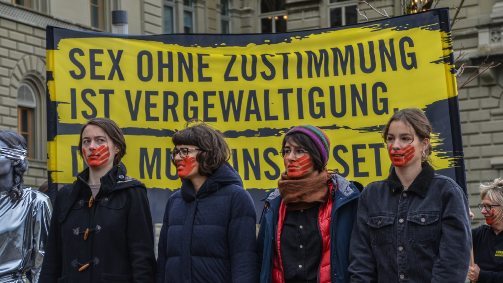 Cuatro mujeres con una huella roja en la boca se paran frente a una pancarta que pide que se actualicen las leyes obsoletas de Suiza sobre violencia sexual