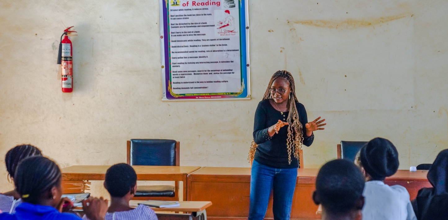 A teacher stands at the front of a classroom as she speaks to a group of students.