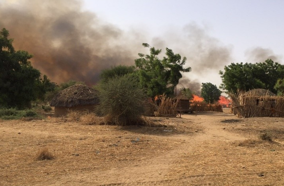 A large fire blazes behind huts and trees, smoke billowing into the sky.