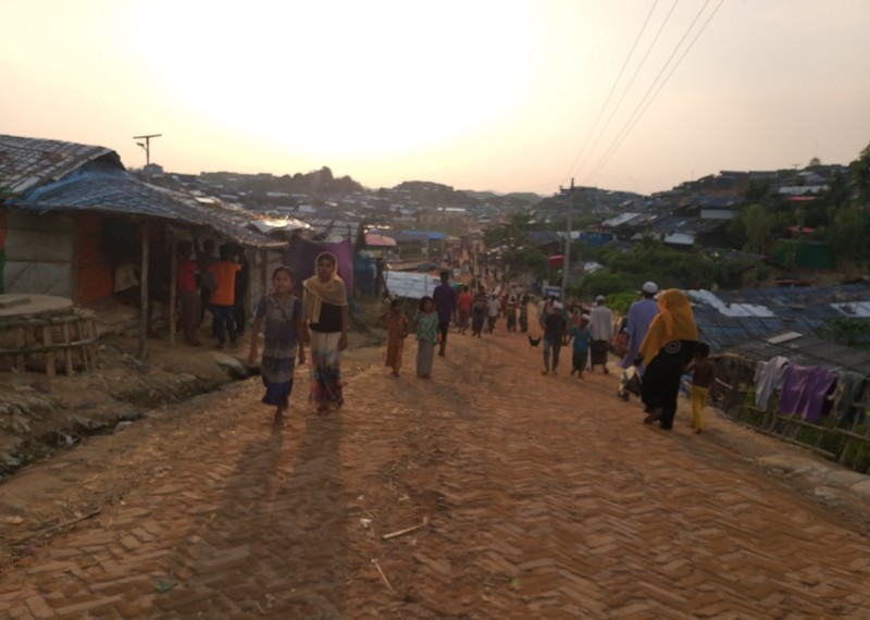 People walk up and down a large street within a refugee camp, the sun setting over the hill in the background.