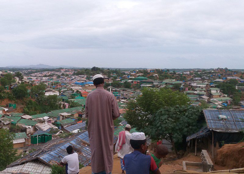 Some people stand with their backs to the camera, looking out onto a large refugee camp. 