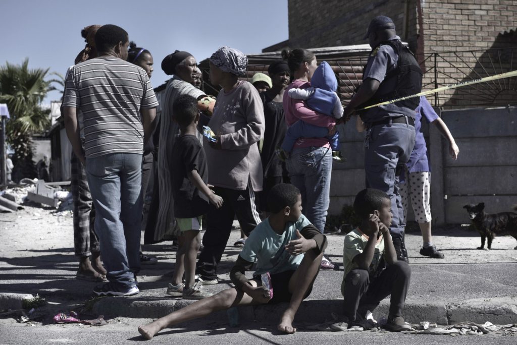 A group of people, adults and children, with expressions that show both curiousity and worry, as a police officer puts up police tape.
