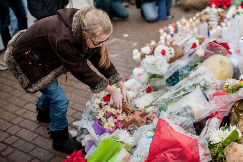 a young person, likely around 9 years old, leans down to place a wooden cross  on a memorial for the victims of the Sandy Hook shooting. The memorial is full of lit candles, bouquets of flowers and stuffed teddy bears.  