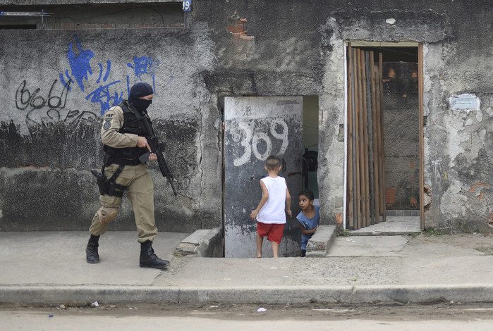 two young boys rush indoors as an armed officer walks down their road carrying a large rifle.