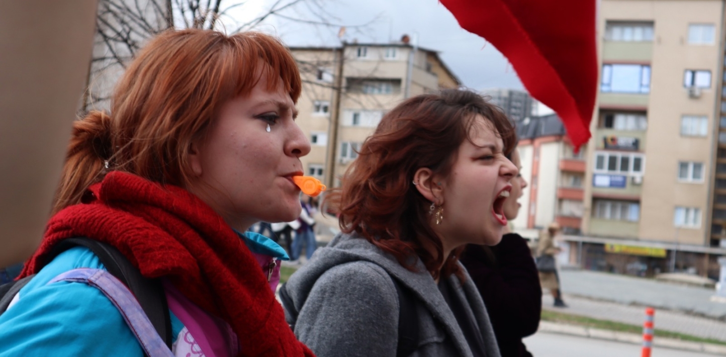 Image shows the profiles of the faces of two women participating in a march in Kosovo against domestic violence. The woman on the left is wearing a red scarf, has a diamante 'tear' below her left eye, and an orange whistle in her mouth. The woman on the right is wearing a grey coat, multiple earrings, and is shouting.