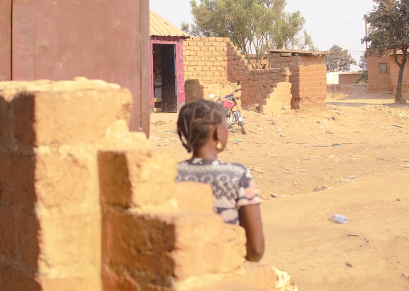 A child stands in amongst buildings, we see the back of her head and shoulders.