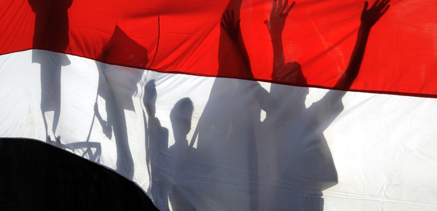 The shadows of protesters hits against the red, white and black national flag of Yemen during a commemoration in 2016 of the 26 September revolution 1962 that established Yemen as a republic.