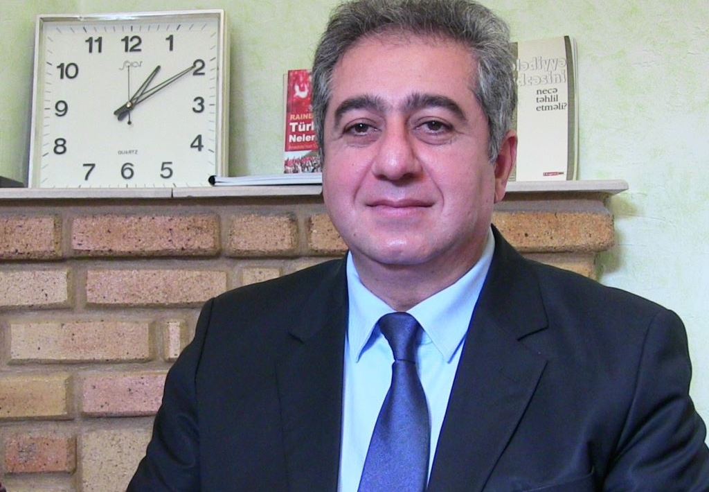 A portrait of Gubad Ibadoghlu. He is looking at the camera while wearing a dark suit and a blue tie on a white shirt. There is a clock in the background of the image.