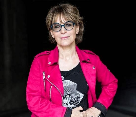 Agnes Callamard is wearing a pink jacket, black t shirt and large blue, round framed glasses. 