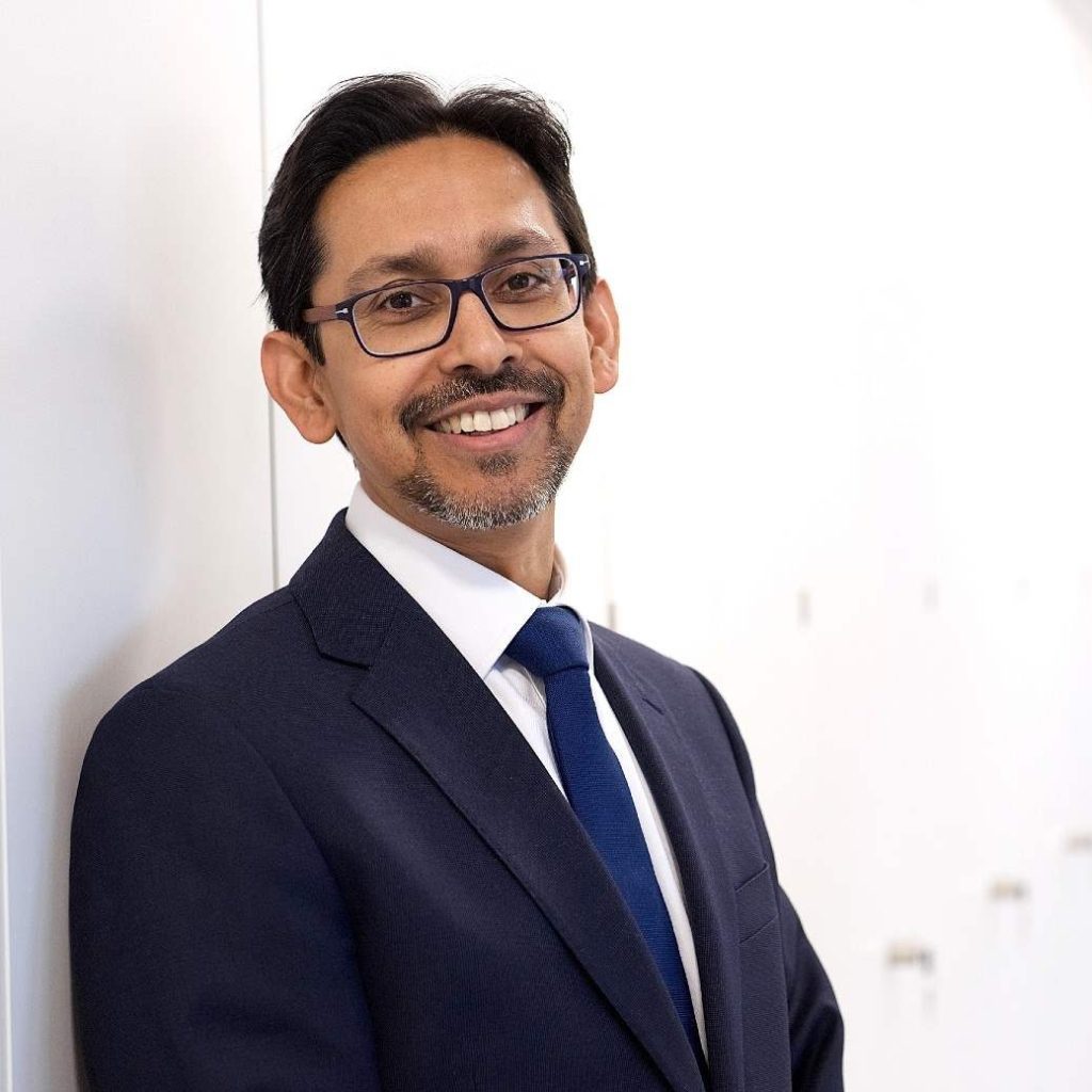Rohan Hewavisenti, standing in front of a white wall. He is wearing glasses, a blue tie, white collared shirt and a jacket.