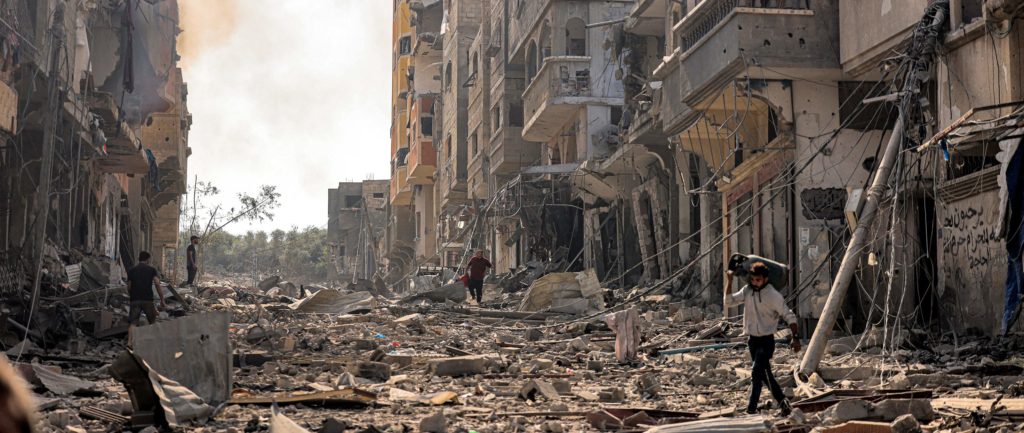 An aftermath of a bombed area in Gaza. Rubble and destroyed buildings everywhere. A man is walking on top of the rubble.