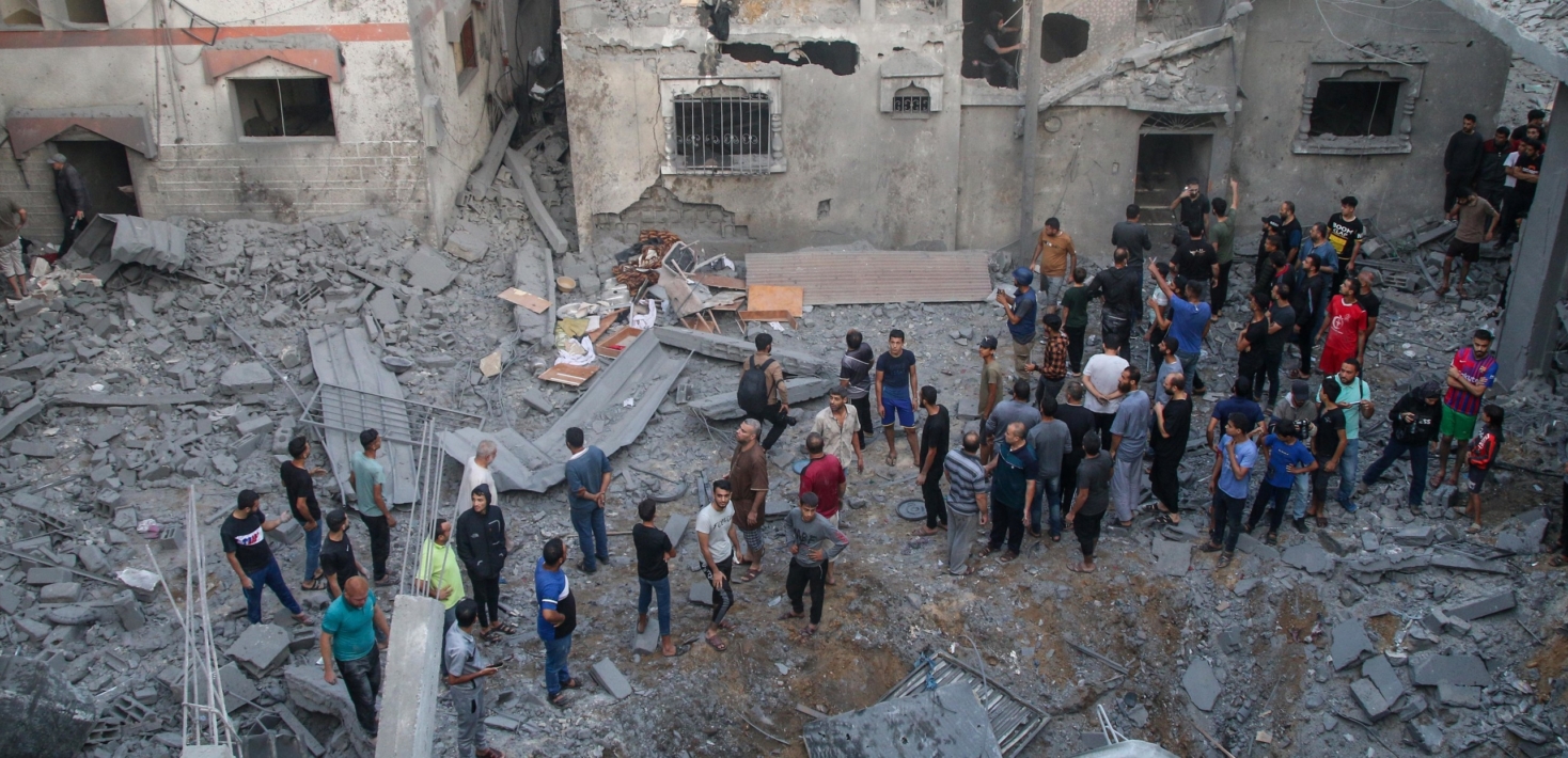 A crowd of people standing amongst rubble, and bombed building.