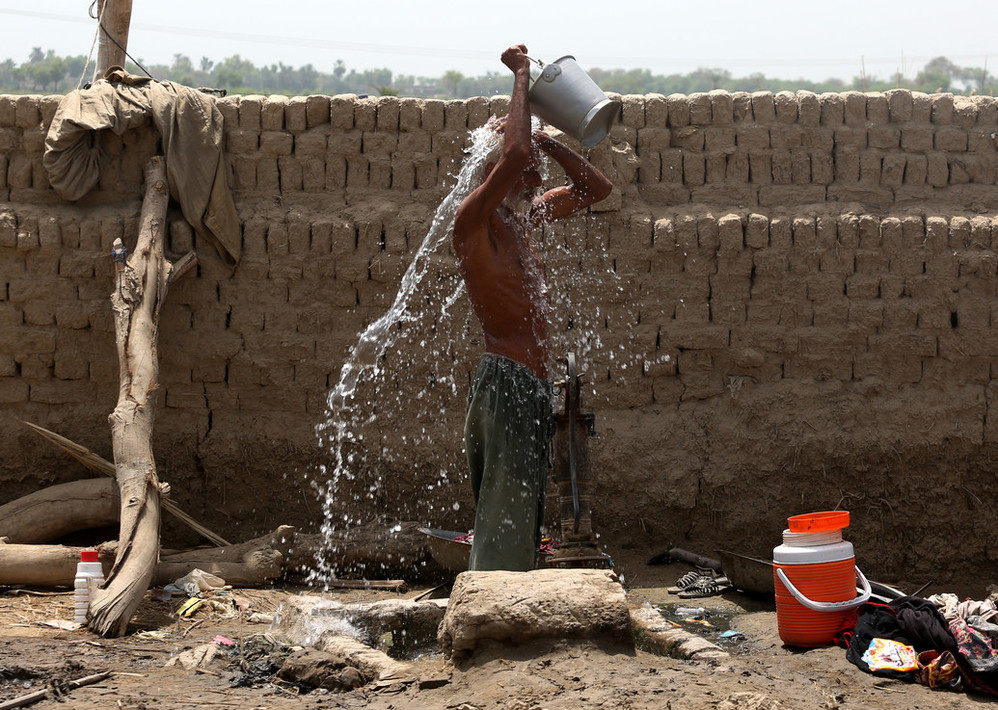 a man is pouring a bucket of water over his head. behind him is a newly set brick wall. the earth around him is dry and dusty. 