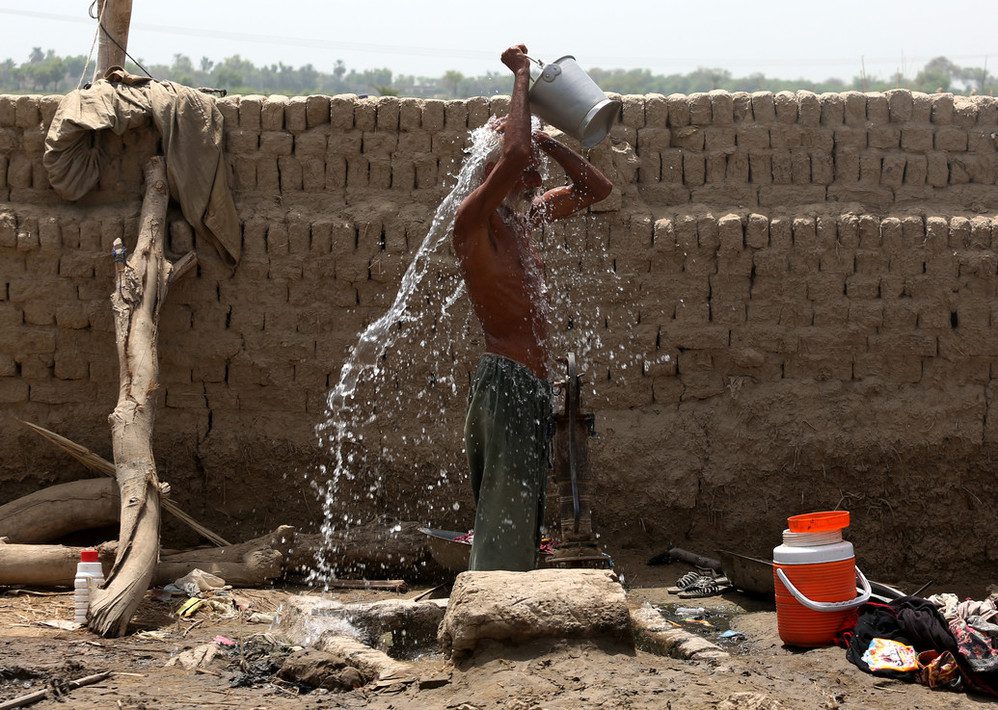 a man is pouring a bucket of water over his head. behind him is a newly set brick wall. the earth around him is dry and dusty.