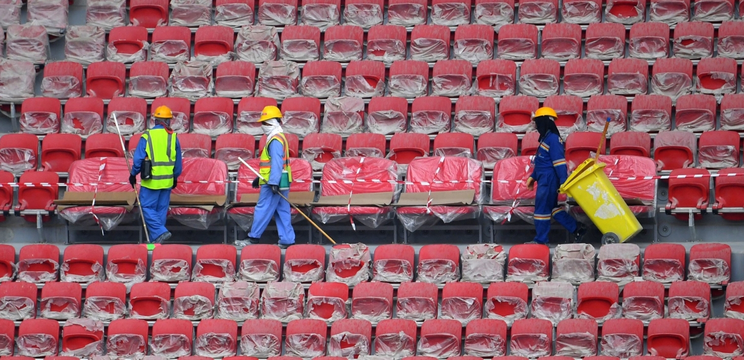 Workers on a walkway in a stadium between a bank of empty red seats