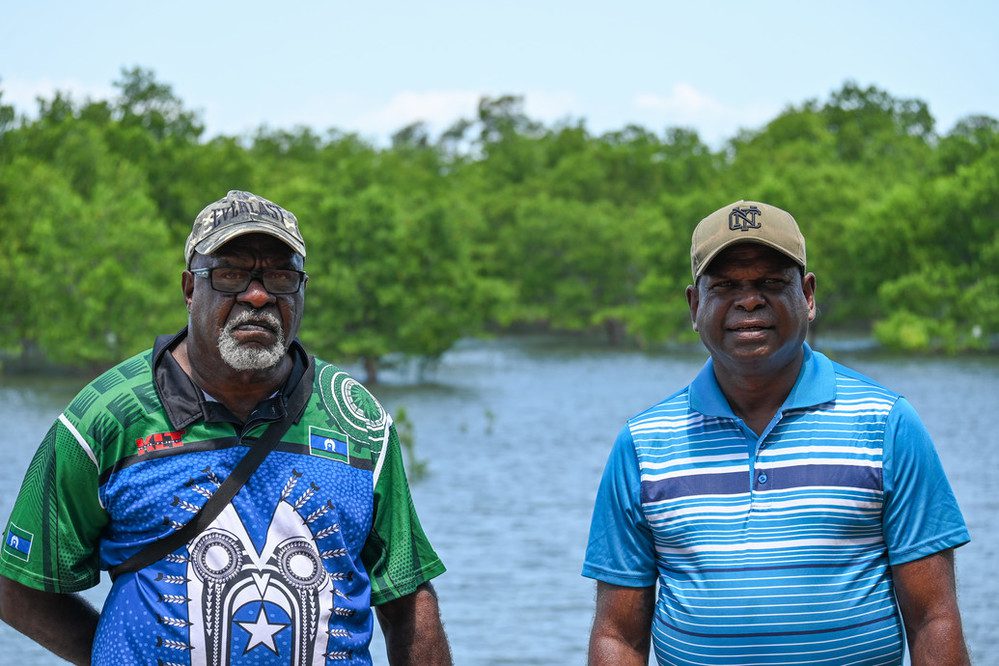 Dos hombres están de pie delante de una masa de agua rodeada de vegetación. Ambos llevan gorras de béisbol y camisetas coloridas. 