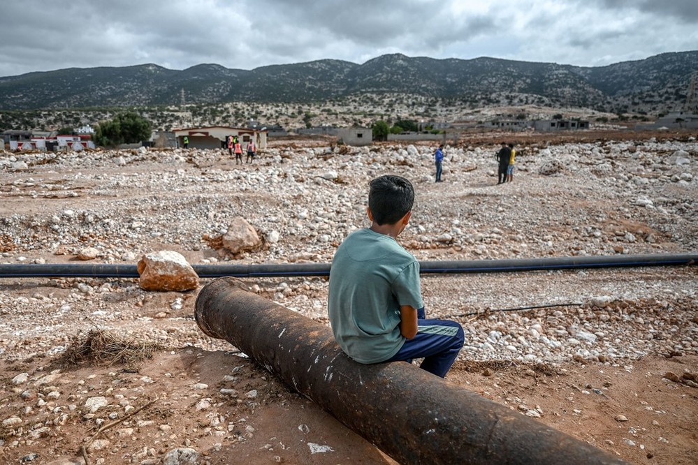a young boy sits on an old rusty pipe, overlooking the rubble of a city impacted by floods