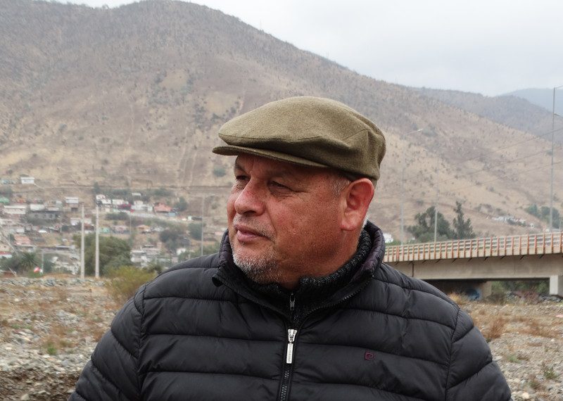 Un hombre con abrigo y gorra mira hacia un lado; al fondo, un puente y una montaña.