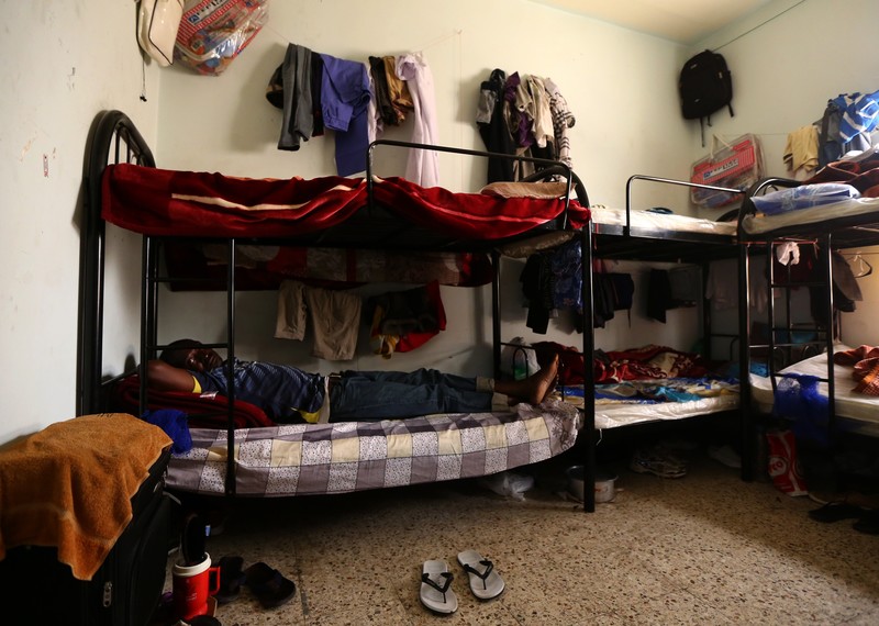 A man lies on a bunkbed in a cramped room, surrounded by clothes. 