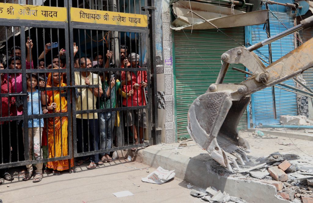 a large group of people stand behind a metal gate and look at a bulldozer. Some of them are taking videos with their phones