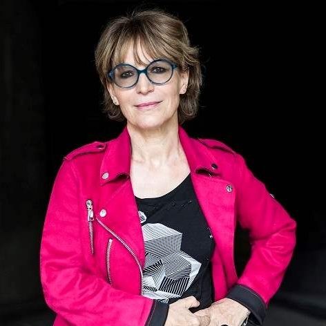 Agnes Callamard is wearing a pink jacket, black t shirt and large blue, round framed glasses. 