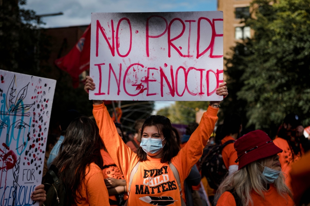 une femme portant un sweat-shirt orange qui arbore le slogan « Every Child Matters » (chaque enfant compte) et tenant une pancarte sur laquelle il est inscrit « No Pride in Genocide » (pas de fierté dans le génocide).