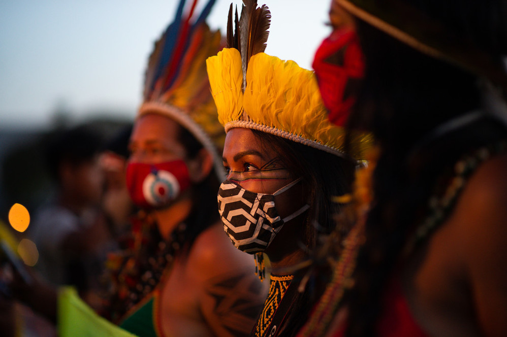 a group of people stand in a line. They are wearing patterned face masks and colourful feathered headdresses. 