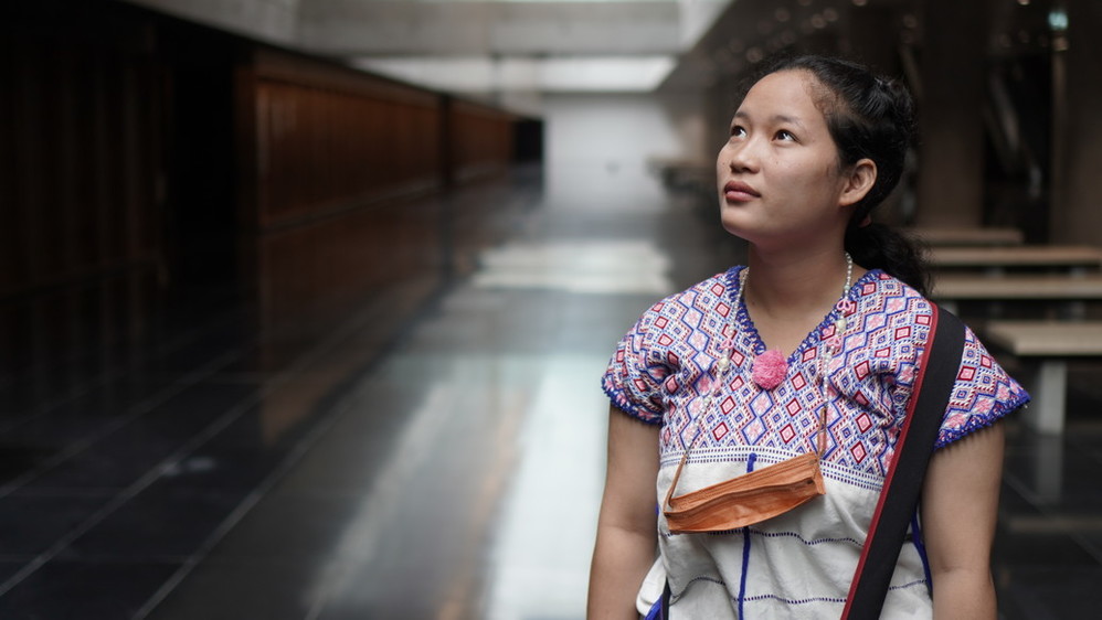 a portrait of Chan Tonnampetch, a young activist from Thailand. She is wearing a pink patterned shirt and a face mask hanging from her neck on a beaded necklace. 