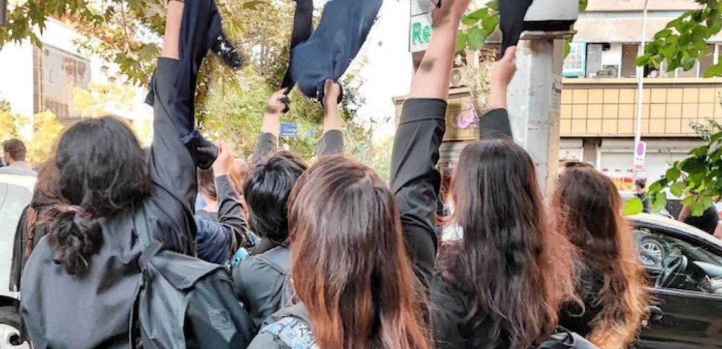 An image of Iranian female students seen with their faces turned away from camera and their brown hair loose taking ff their black headcovers and raising them in the air in a sign of defiance.