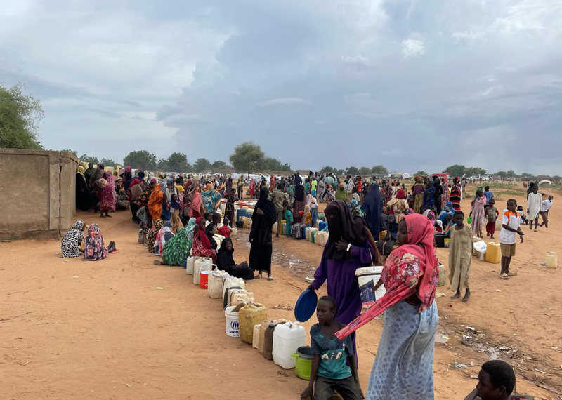 Женщины и дети стоят в длинной очереди с емкостями для воды. 