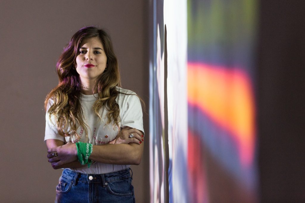 a portrait of Sofia, who is posing with her arms crossed. She has a green bandana tied around her wrist. 