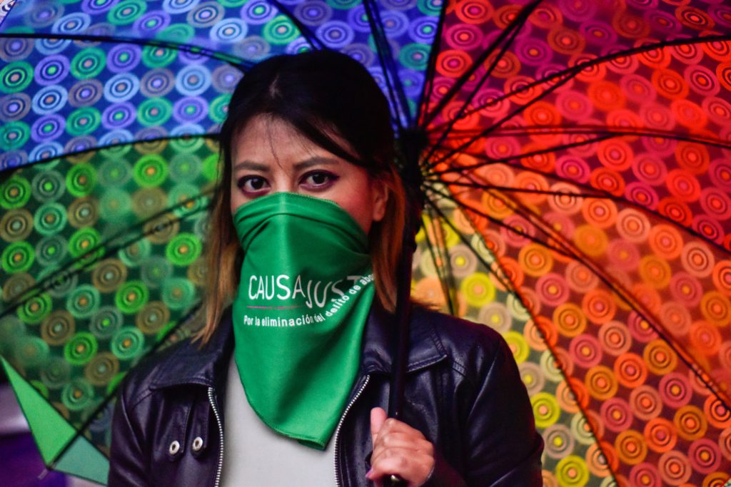 someone standing under a rainbow umbrella and wearing a green bandana over the lower part of their face