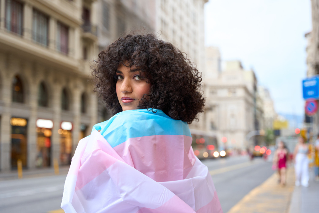 a trans person posing with the pink, white and blue trans flag wrapped around their shoulders.