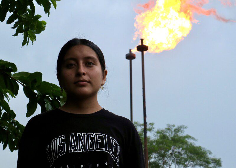 A young woman stands in front of a gas flare.