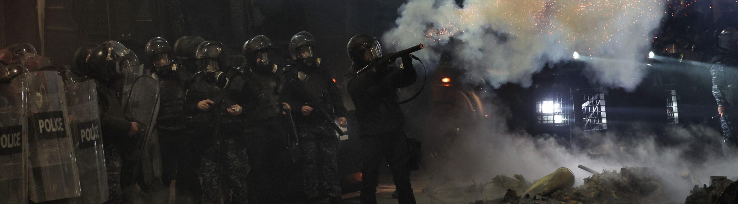 The image shows a line of riot police in full gear, with shields and helmets, amidst heavy smoke and sparks, as one officer fires a projectile to disperse protesters in a tense nighttime scene