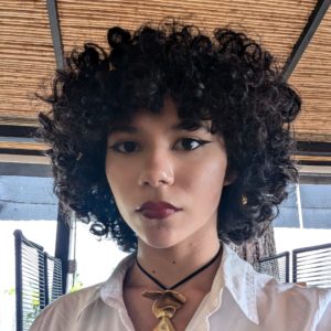 Eunice mira a la cámara con expresión firme y seria. Viste camisa blanca y lleva un colgante dorado grande.