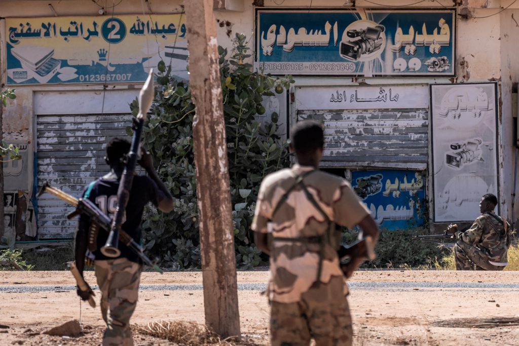 Three armed men take positions on a street in front of shuttered storefronts. Two of them have their backs to us. The man on the left, stepping into the midday sunlight to cross the street, holds a sheathed dagger in his left hand, and slings a rocket propelled grenade launcher and an AK-47 over his back. He wears camouflage pants and a football shirt. In the middle, a man, also with his back to us, wears full military fatigues cradling an AK-47. Across the street, squatting on the curb in the shade of the storefronts, is a third man in fatigues, half his face visible, with a machine gun on his left knee, pointing down in the direction where he is looking. Besides these three men, the street is empty.