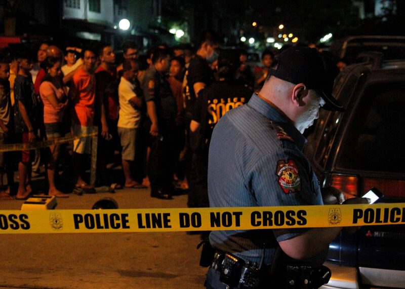 A photo at night of a policemen behind a police line waiting at the scene of an anti-drug police operation, with bystanders watching.
